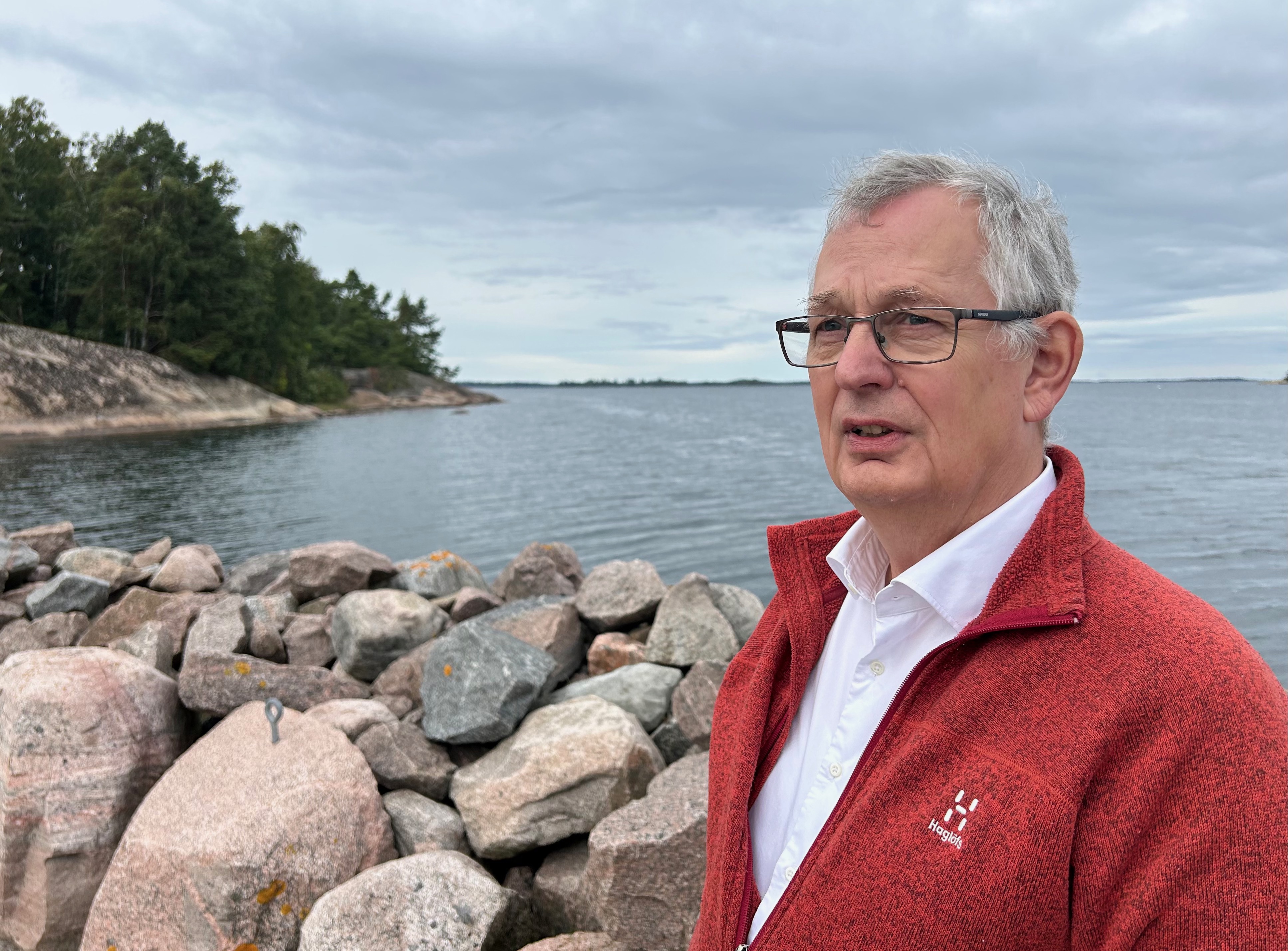 Mann i rød jakke med vann og stener i bakgrunn.foto